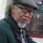Portrait of an older man with glasses and a gray beard wearing a checkered flat cap green jacket and purple shirt indoors